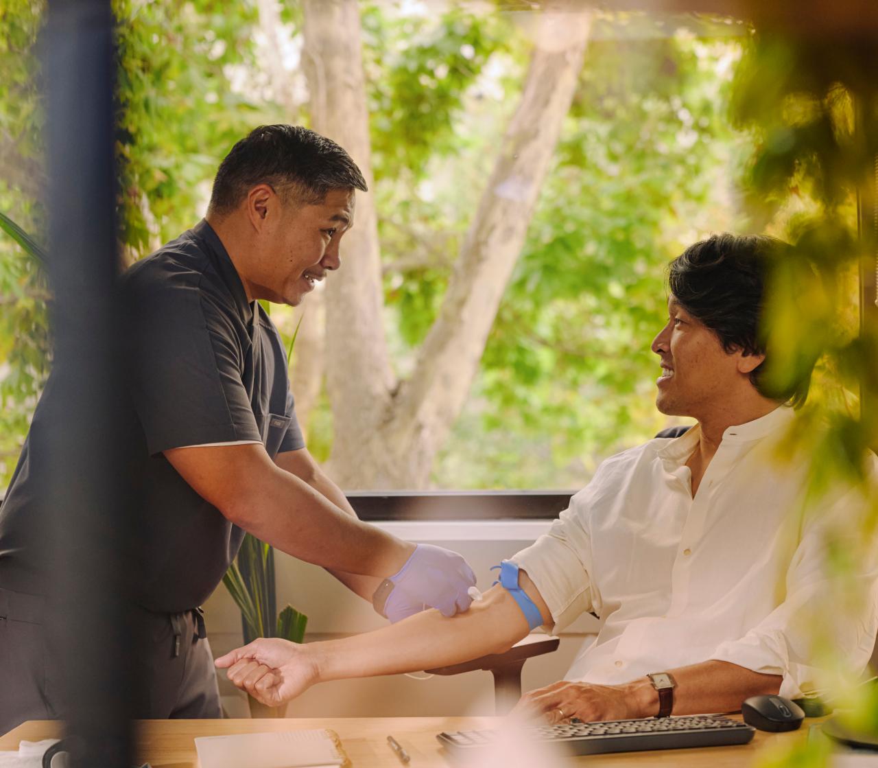 A gloved phlebotomist prepares to draw blood from a smiling man's arm at his office desk, with greenery visible through the window behind them.