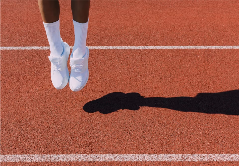 Person's feet in white sneakers above a red running track with a strong shadow.