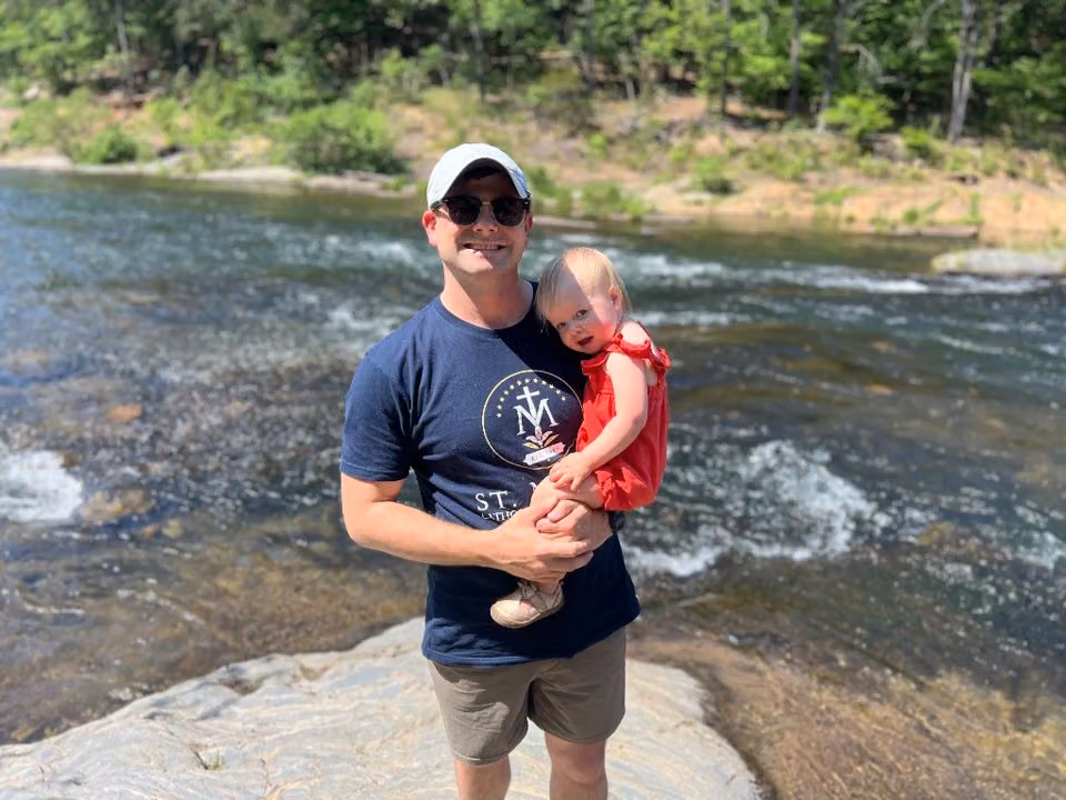Tanner holding his young daughter outdoors by a river.