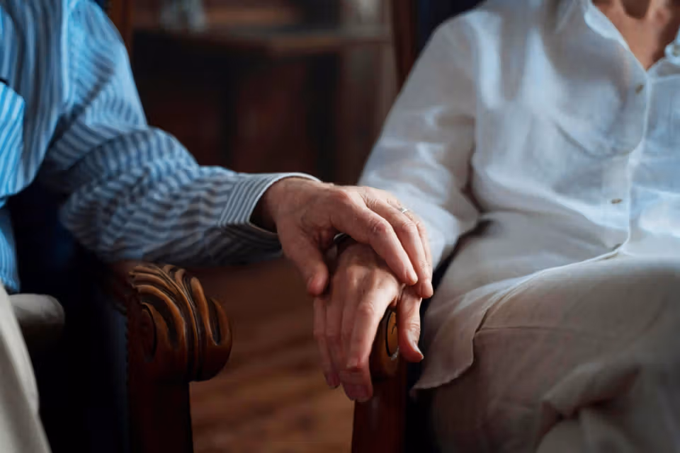 Close-up of two older adults holding hands while seated indoors.