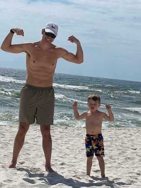 Travis flexing on a beach beside a young child near the shoreline.