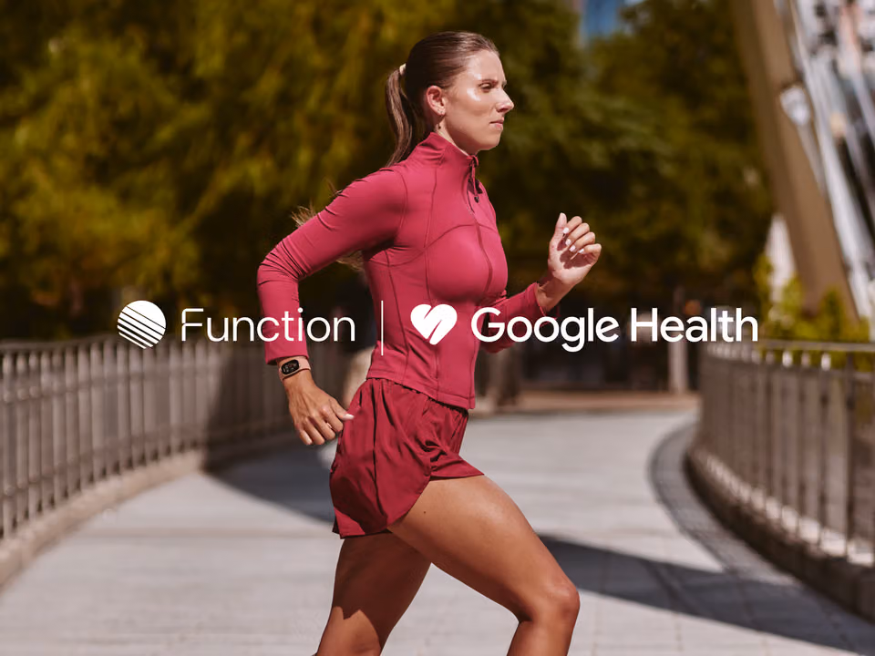 Woman in a magenta jacket and shorts running across a bridge, wearing a fitness tracker, with the Function and Google Health logos overlaid in white.