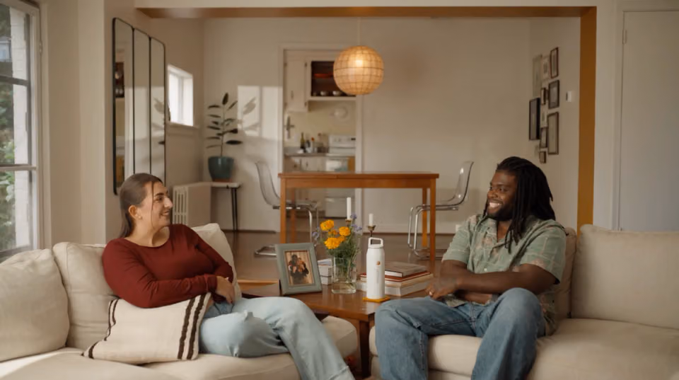 Sara and Tré smiling at each other while seated in their living room.