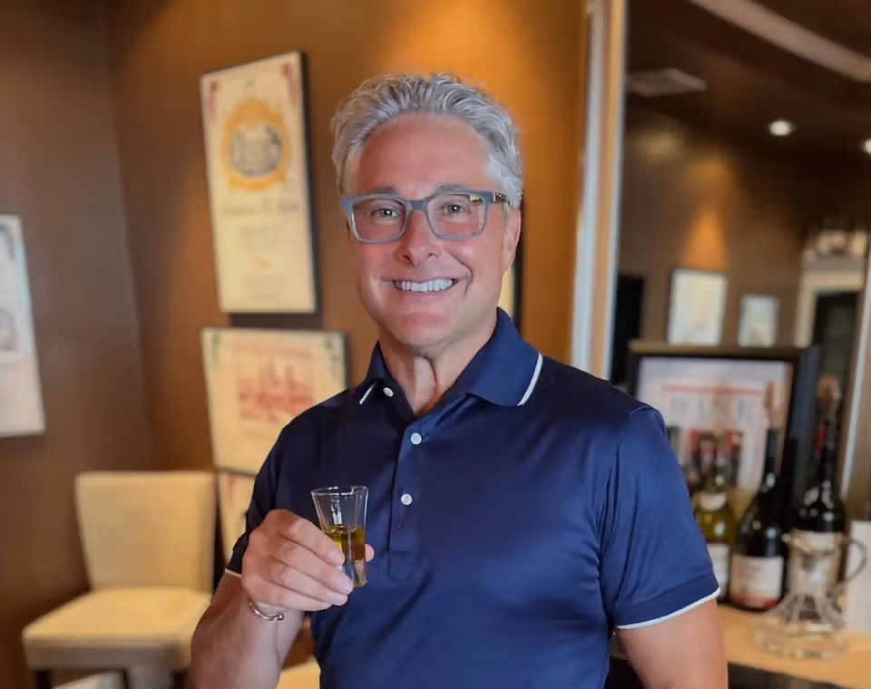 Ralph holding a small glass indoors and smiling at the camera.