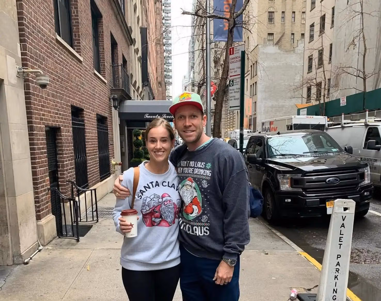 Holly and a man standing together on a city sidewalk in holiday sweatshirts.