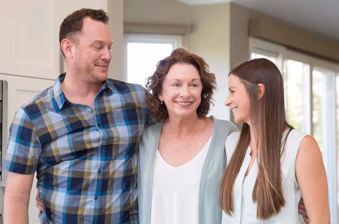 Three people standing together indoors and smiling in a bright kitchen.